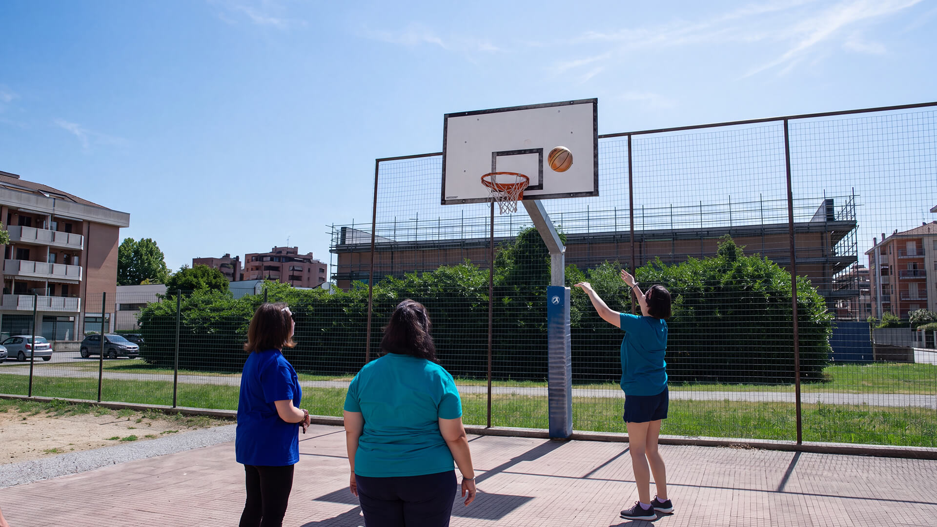 Tre persone su un campetto all'aperto guardano il pallone entrare nel canestro da basket