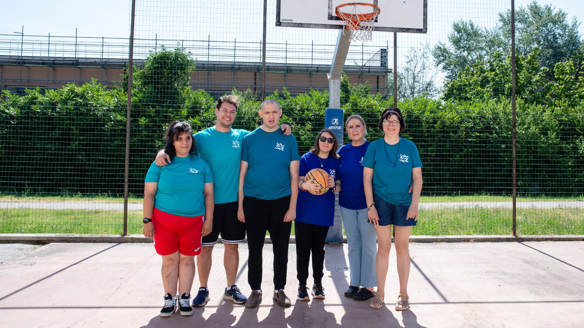 Sei persone posano sorridenti sul campo da basket all'aperto, una tiene il pallone tra le mani