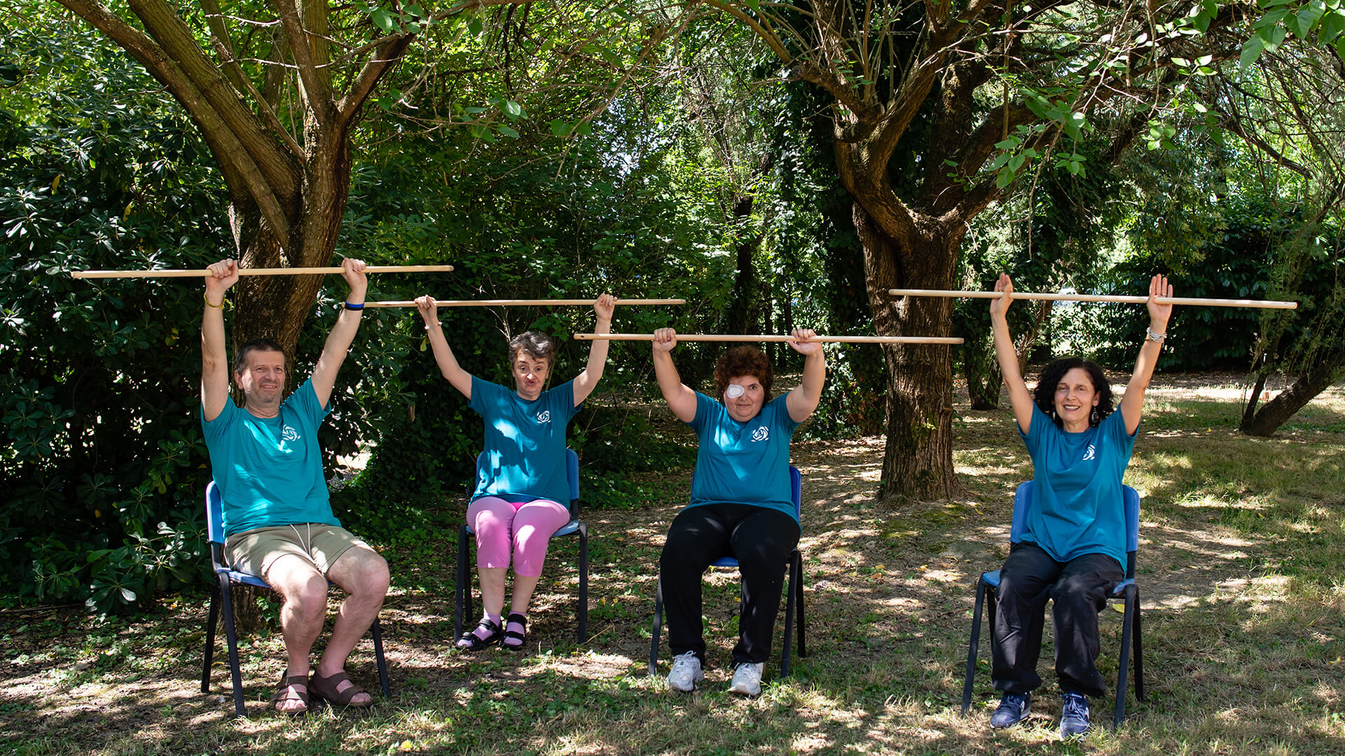 Quattro persone sedute all'aperto sollevano bastoni di legno durante un esercizio di ginnastica dolce